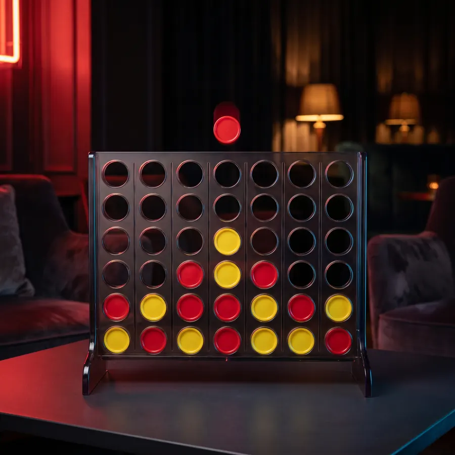 A vertical grid game standing on a dark table, part-filled with red and yellow discs and one held above a slot.