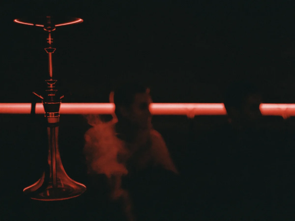 A hookah silhouetted against a red neon strip in a dark lounge, smoke drifting past two seated guests.