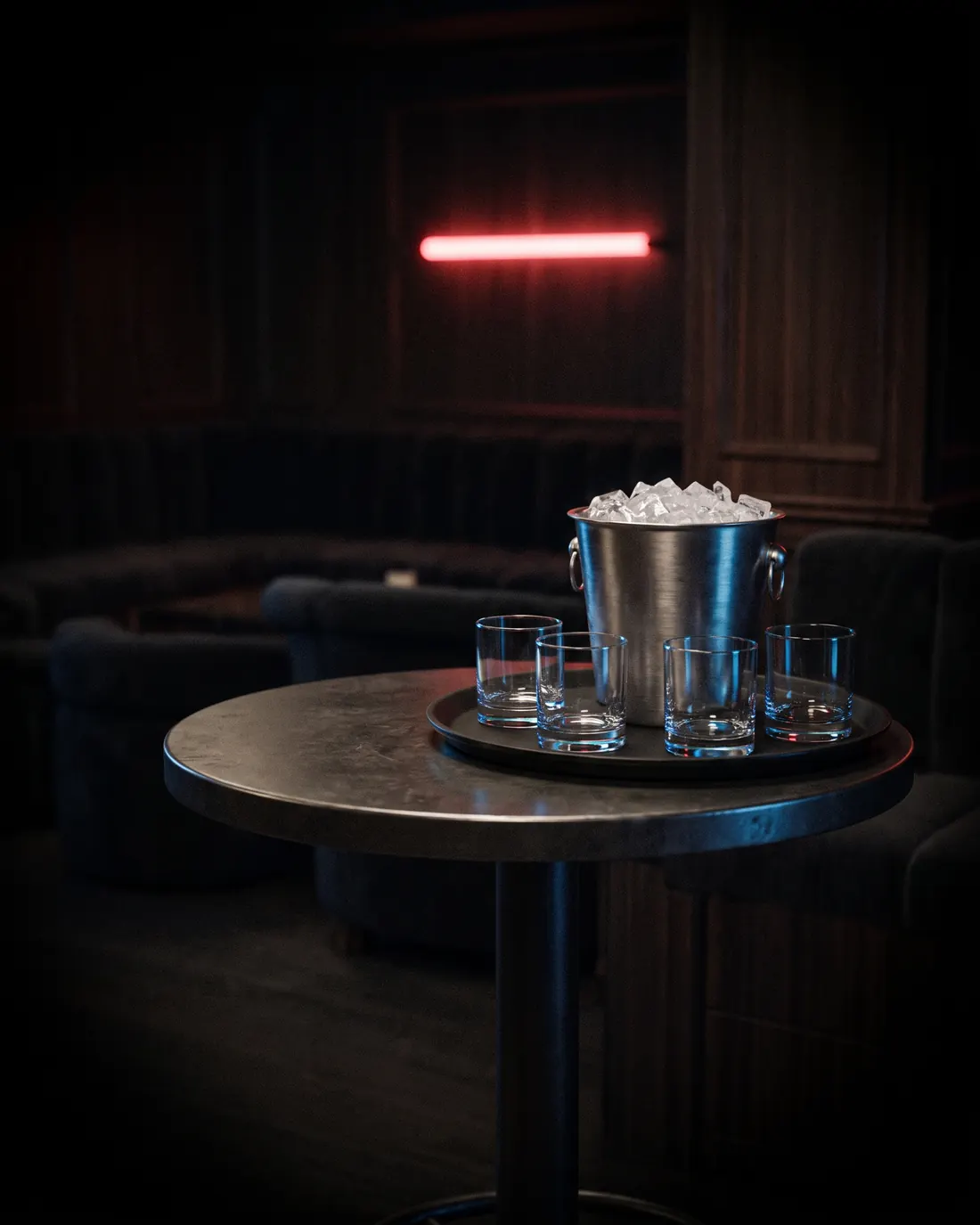 A high-top table in the dark lounge set with a steel ice bucket and four empty tumblers, one red neon line on the wall behind.
